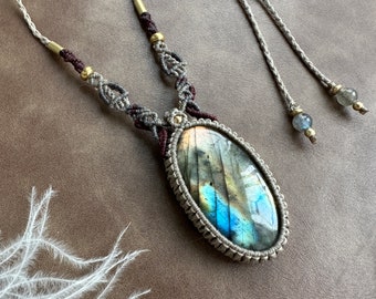 Puede incluir: Un collar de macramé plateado y marrón con un gran colgante ovalado de labradorita. El colgante tiene colores iridiscentes azules, verdes y dorados.