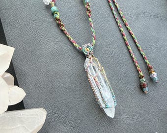 May include: A clear, pointed quartz crystal pendant encased in a woven macrame cage of green, pink, and gold threads. The necklace features a braided cord adorned with small, multi-colored glass beads and additional clear crystal accents.