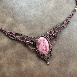 May include: A handmade macrame necklace featuring a large, oval pink gemstone pendant. The necklace is crafted with dark brown cord, accented with small gold-colored beads. The intricate macrame design surrounds the gemstone, creating a unique and stylish accessory.