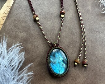 Puede incluir: Un collar con un colgante de labradorita azul en un cordón marrón y verde. El cordón tiene cuentas de madera y está atado con un nudo macramé.