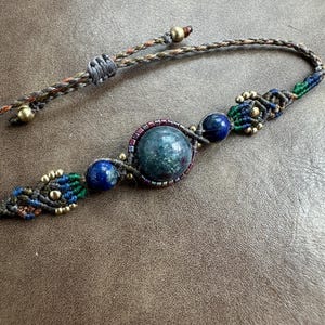 May include: Macrame bracelet featuring a large, round, teal-colored stone as the centerpiece, flanked by two smaller blue stones. The macrame work incorporates blue, green, and brown threads, accented with small gold-colored beads. The adjustable closure is made of braided threads.