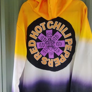 May include: A hooded sweatshirt with a gradient design, transitioning from yellow to white, then to purple and black. The back features a circular graphic with the words "HOT CHILI PEPPERS" and a purple face design.