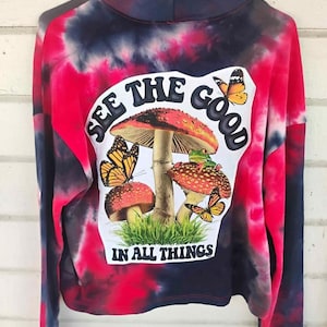 May include: A tie-dye sweatshirt in shades of red, pink, and navy blue. The front features a graphic of mushrooms, butterflies, and the text "SEE THE GOOD IN ALL THINGS". The long-sleeved top has a high neckline.