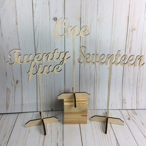 May include: Three wooden table number signs with the numbers "One", "Twenty-five", and "Seventeen" written in a cursive font. The signs are attached to wooden stands with a cross-shaped base.