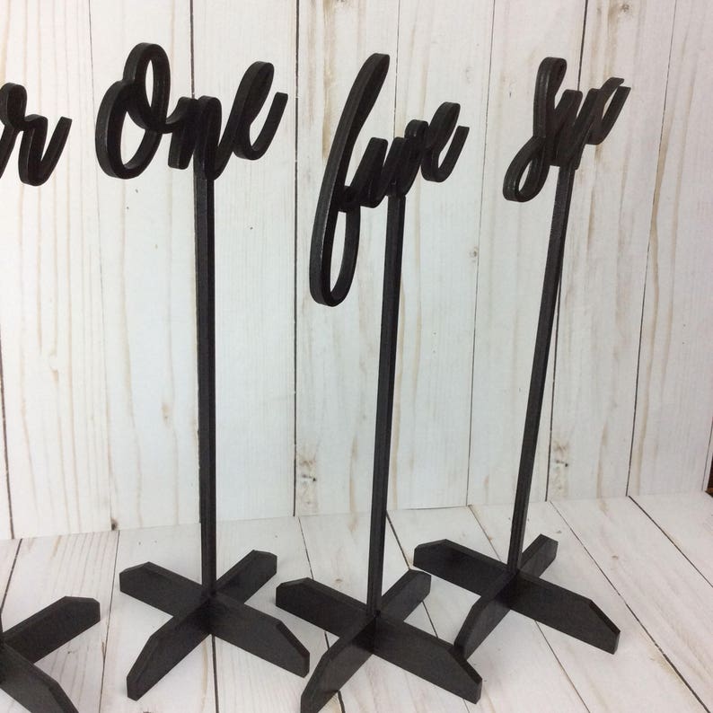 May include: Four black wooden table number holders with the numbers "one", "four", and "six" displayed. The numbers are cut out of the wood and are in a cursive font. The holders have a simple cross-shaped base.