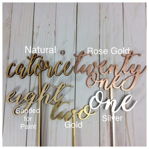May include: Four wooden numbers with different finishes: natural, rose gold, gold, and silver. The numbers are "fourteen", "twenty one", "eight", and "two". The numbers are on sticks and are sanded for paint.