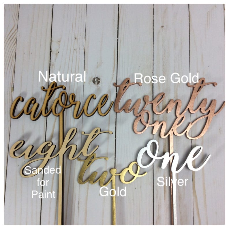 May include: Four wooden numbers with different finishes: natural, rose gold, gold, and silver. The numbers are "fourteen", "twenty one", "eight", and "two". The numbers are on sticks and are sanded for paint.