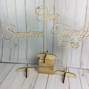 May include: Three wooden table number signs with the numbers "One", "Seventeen", and "Twenty One" written in a cursive font. The signs are on wooden stands.