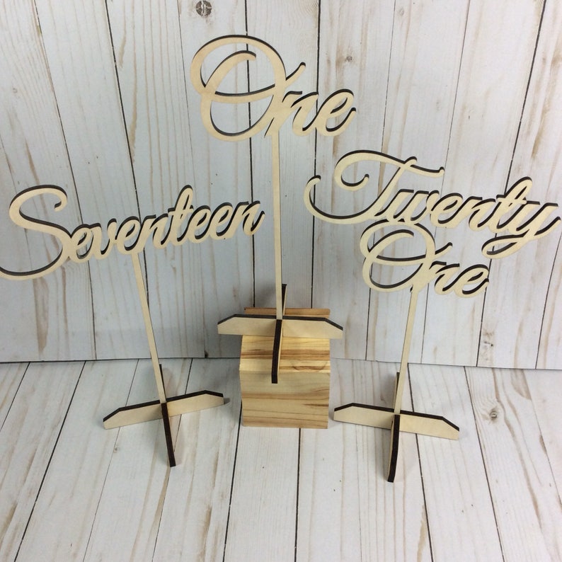 May include: Three wooden table numbers with cursive lettering. The numbers are "One", "Seventeen", and "Twenty One". Each number is attached to a wooden stand.