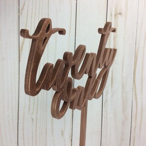 May include: A wooden sign with the words "Twenty One" written in a cursive font. The sign is painted a copper color and is mounted on a wooden stick.