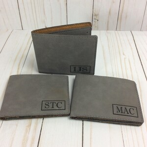 May include: Three gray leather wallets with personalized initials. The wallets have a brown leather interior and are laid out on a wooden surface.