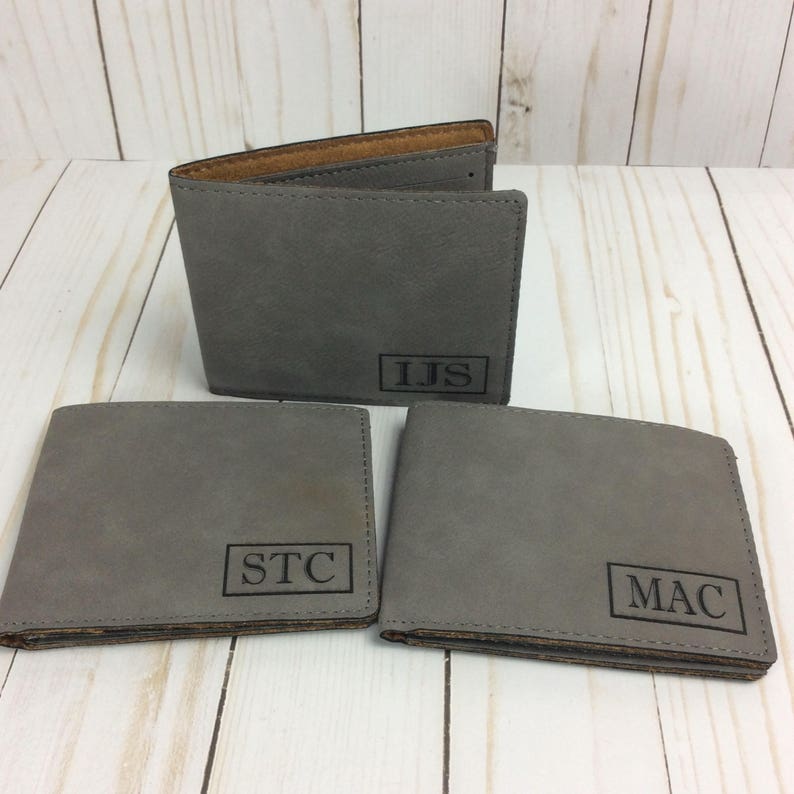 May include: Three gray leather wallets with personalized initials. The wallets have a brown leather interior and are laid out on a wooden surface.