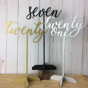 May include: Three table number signs, one gold, one black, and one silver, with the numbers "twenty", "seven", and "twenty one" written in a cursive font.