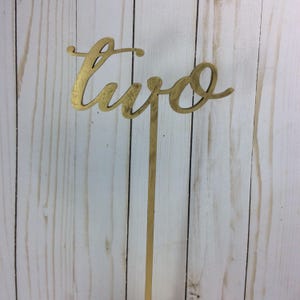 May include: Gold table number with the word "two" written in a cursive script. The number is attached to a gold metal stick.