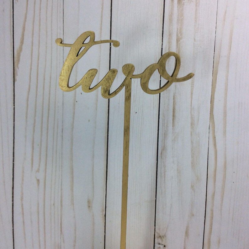 May include: Gold table number with the word "two" written in a cursive script. The number is attached to a gold metal stick.