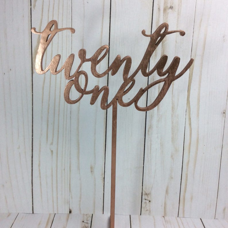 May include: Rose gold colored wooden table number with the text "twenty one" in a cursive font.