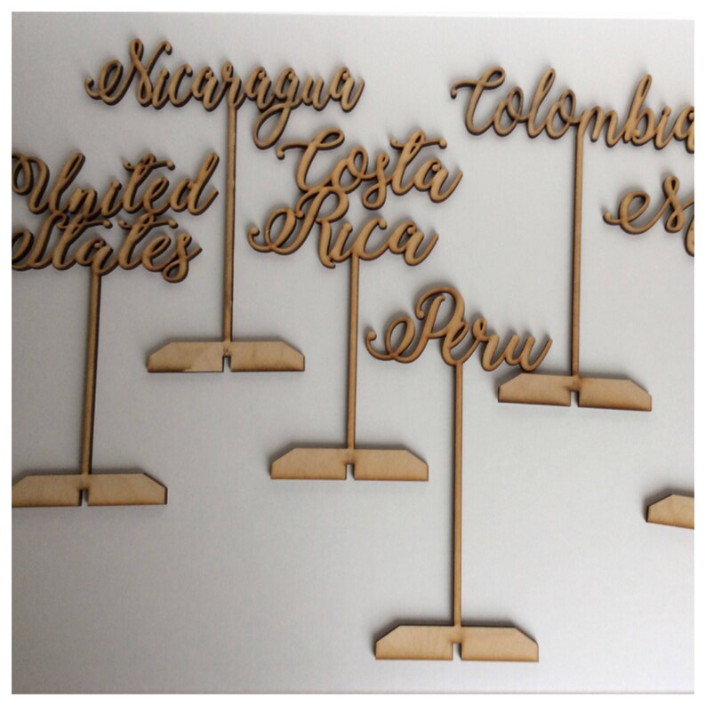 May include: Wooden table number holders with the names of countries written in cursive. The countries are Nicaragua, Costa Rica, United States, Colombia, Peru.