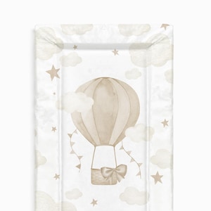 Könnte beinhalten: Eine beige Wickelunterlage mit einem Aquarell-Design aus einem Heißluftballon, Wolken und Sternen. Der Ballon hat einen braunen Korb mit einer Schleife.