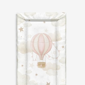 Könnte beinhalten: Eine rechteckige Wickelauflage mit einem skurrilen Design. Die Auflage zeigt einen rosa-weißen Heißluftballon, umgeben von beigen Wolken und Sternen auf weißem Hintergrund. Die Ränder sind in einem weichen Off-White gehalten.