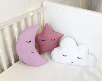 May include: Three plush pillows shaped like a crescent moon, a star, and a cloud. The pillows are all pink and white and have black embroidered eyes and mouths. The pillows are arranged on a white crib mattress.