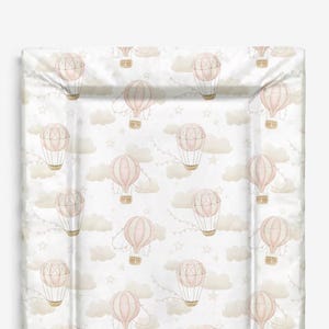 Puede incluir: Un cambiador rectangular con un patrón repetido de globos aerostáticos rosas, nubes y estrellas sobre un fondo blanco. El cambiador tiene un borde elevado para mayor seguridad y está diseñado para el cuidado infantil.