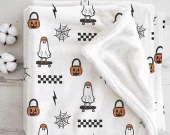 May include: White Halloween-themed blanket with a repeating pattern of ghosts on skateboards, jack-o'-lanterns, spiderwebs, lightning bolts, and checkered squares. The blanket is folded, showing a soft, plush texture.