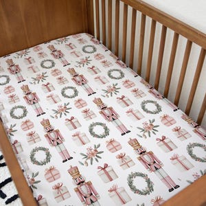 Könnte beinhalten: Ein weißes Spannbettlaken für das Kinderbett mit einem sich wiederholenden Muster aus rosa Nussknackern, verpackten Geschenken, Blumensträußen und grünen Kränzen. Das Laken befindet sich in einem Holzbett.