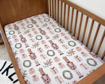 May include: A white fitted crib sheet with a repeating pattern of pink nutcrackers, wrapped gifts, floral bouquets, and green wreaths. The sheet is inside a wooden crib.