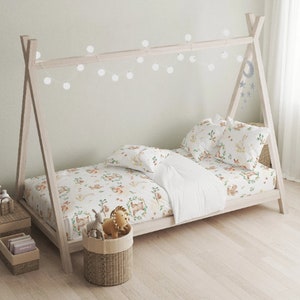 May include: A light wood teepee-style bed frame with white bedding and woodland-themed sheets and pillowcases. The bedding features a pattern of animals and foliage. A string of white globe lights hangs above the bed. A woven basket holds stuffed toys.