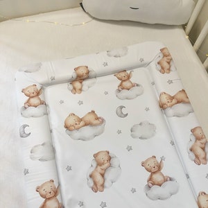 Könnte beinhalten: Eine weiße Wickelauflage mit einem Muster aus braunen Teddybären auf Wolken, grauen Sternen und Monden. Ein weißes wolkenförmiges Kissen mit geschlossenen Augen liegt über der Auflage. Die Auflage liegt in einem weißen Kinderbett.