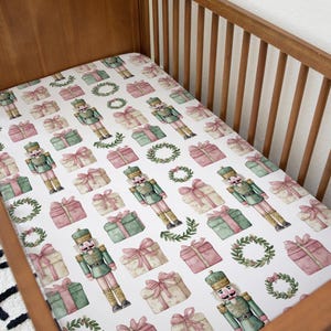 May include: A white cot sheet featuring a repeating pattern of watercolour nutcrackers, wrapped gifts, and wreaths in green, pink, and gold. The sheet is inside a wooden cot.