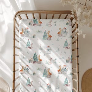 Könnte beinhalten: Weißes Spannbettlaken für ein Babybett mit einem sich wiederholenden Muster aus einem Schneemann, einem Fuchs, einem Kaninchen, einem Igel und einem Weihnachtsbaum. Das Muster ist in Grün-, Braun-, Rot- und Weißtönen gehalten.