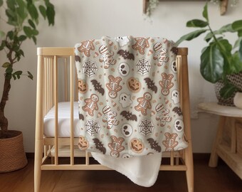 Op de afbeelding: Een beige Halloween-deken gedrapeerd over een houten wieg. De deken heeft een herhalend patroon van peperkoeken skeletten, mummies, vleermuizen, spinnenwebben, schedels, geesten en pompoenen in bruin en oranje.