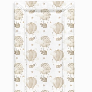 Puede incluir: Una cambiador blanco con un patrón de globos aerostáticos beige y marrón. Los globos tienen un lazo marrón y están rodeados de nubes blancas y estrellas doradas.