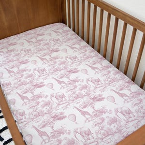 May include: A fitted cot sheet with a pink and white toile pattern featuring giraffes, elephants, and hot air balloons. The sheet is inside a wooden cot.