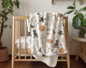 Op de afbeelding: Een witte Halloween-thema babydeken met een zachte, pluche textuur. De deken heeft oranje pompoenen, zwarte katten, vleermuizen, geesten en andere griezelige illustraties. De deken is over een houten wieg gedrapeerd.