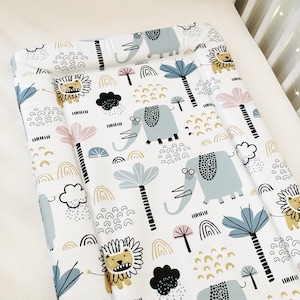 May include: A white changing mat with a colourful pattern of elephants, lions, palm trees, and rainbows. The mat is made of soft, comfortable fabric and is ideal for changing babies.