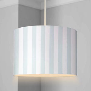 Peut inclure: Abat-jour cylindrique à rayures verticales bleu clair et blanches. L'abat-jour est suspendu au plafond par un cordon fin et blanc cassé. La lumière est allumée, éclairant le bas de l'abat-jour.