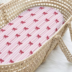 Puede incluir: Una sábana de cuna para bebé con rayas rosas y blancas y un patrón de lazos rojos. La sábana está dentro de una cuna de mimbre tejida. La cuna tiene asas y está sobre una superficie blanca y esponjosa.