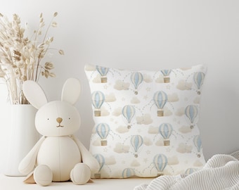 May include: A white throw pillow with a blue and brown hot air balloon pattern. The pillow is on a white surface with a white bunny plush toy in front of it.