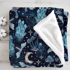 Può includere: Una coperta blu scuro con un design celeste con cristalli, funghi, stelle e una luna crescente. La coperta ha un lato inferiore bianco e soffice. Il design è nei toni del blu e del bianco.