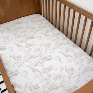 May include: A white cot sheet with a beige safari animal print featuring giraffes, elephants, and hot air balloons.