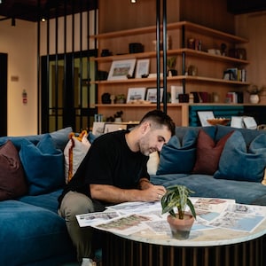 May include: A person sitting on a blue couch, working on a table with a plant and papers spread out. The person is wearing a black shirt and khaki pants.