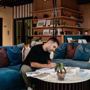 May include: A person sitting on a blue couch, working on a table with a plant and papers spread out. The person is wearing a black shirt and khaki pants.