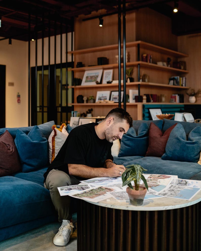 May include: A person sitting on a blue couch, working on a table with a plant and papers spread out. The person is wearing a black shirt and khaki pants.