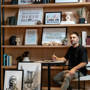 Pu&ograve; includere: Un uomo &egrave; seduto su una sedia di fronte a una libreria con diverse opere d'arte incorniciate e libri. Le opere d'art includono illustrazioni in bianco e nero di paesaggi urbani e mappe. L'uomo indossa una camicia nera, pantaloni kaki e scarpe da ginnastica bianche. Sta tenendo una matita e un quaderno.