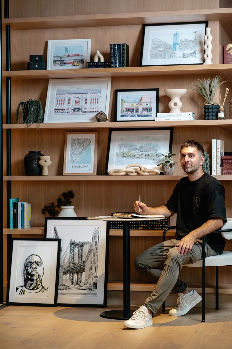 Pu&ograve; includere: Un uomo &egrave; seduto su una sedia di fronte a una libreria con diverse opere d'arte incorniciate e libri. Le opere d'art includono illustrazioni in bianco e nero di paesaggi urbani e mappe. L'uomo indossa una camicia nera, pantaloni kaki e scarpe da ginnastica bianche. Sta tenendo una matita e un quaderno.