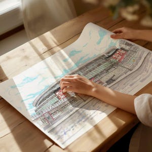 May include: A hand traces the outline of a detailed drawing of a stadium, with the word "Arsenal" visible on the side. The drawing is on a white sheet of paper, with a light blue sky and clouds in the background.