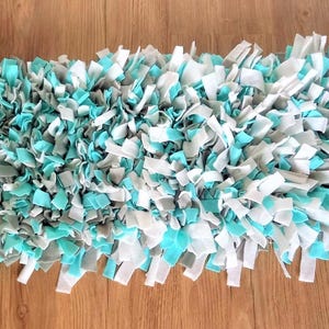 Könnte beinhalten: Ein rechteckiger Flickenteppich mit zotteliger Textur. Der Teppich besteht aus Stoffstreifen in Weiß-, Grau- und Türkistönen. Der Teppich liegt auf einer Holzoberfläche.