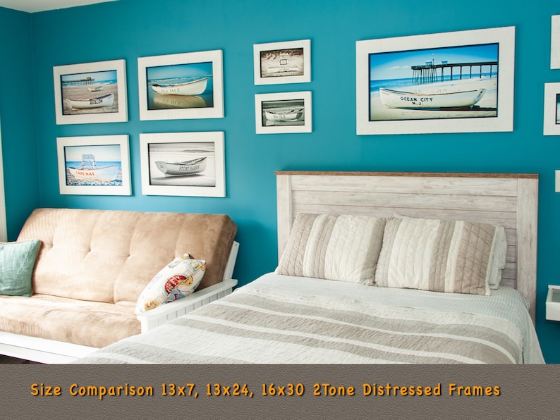 May include: A selection of framed art prints with a coastal theme, set in white distressed frames. The prints show boats on sandy beaches and a pier. The frames are hung on a teal wall above a bed and futon. One print displays the text "Ocean City N.J.".